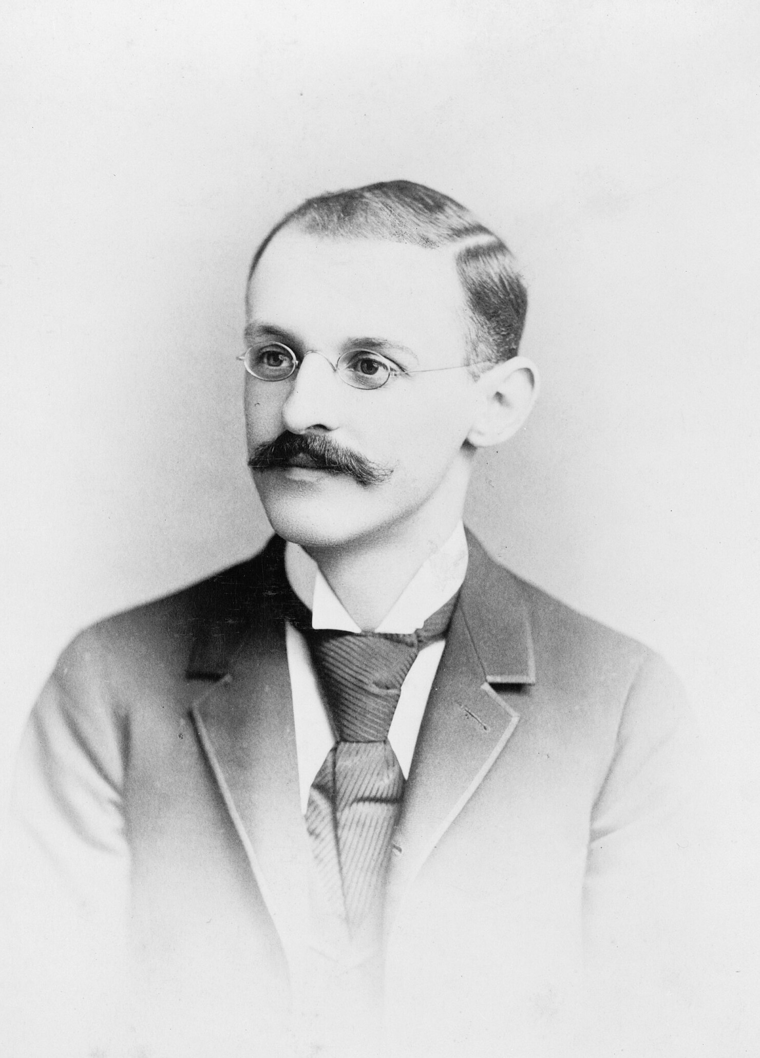 Retrato antiguo en blanco y negro de Abraham Flexner joven, con bigote, gafas pequeñas, traje y corbata, mirando hacia la izquierda
