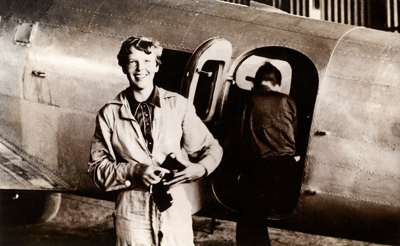 Fotografía antigua de Amelia Earhart sonriente, con mono de vuelo, junto a la puerta de su avión metálico, por la que sube un hombre