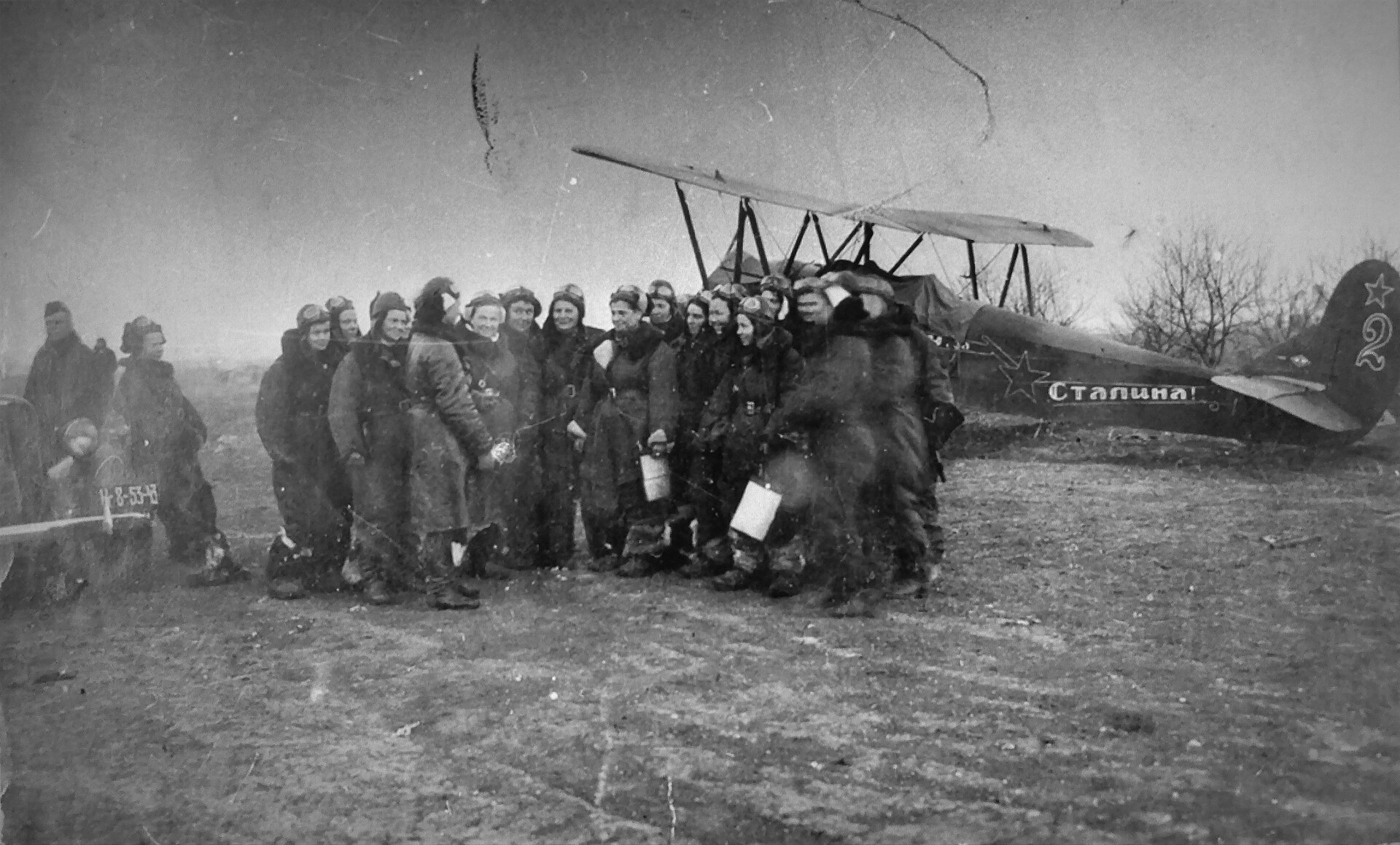 Fotografía en blanco y negro de un grupo de mujeres jóvenes con uniforme de aviadora delante de un pequeño avión biplano