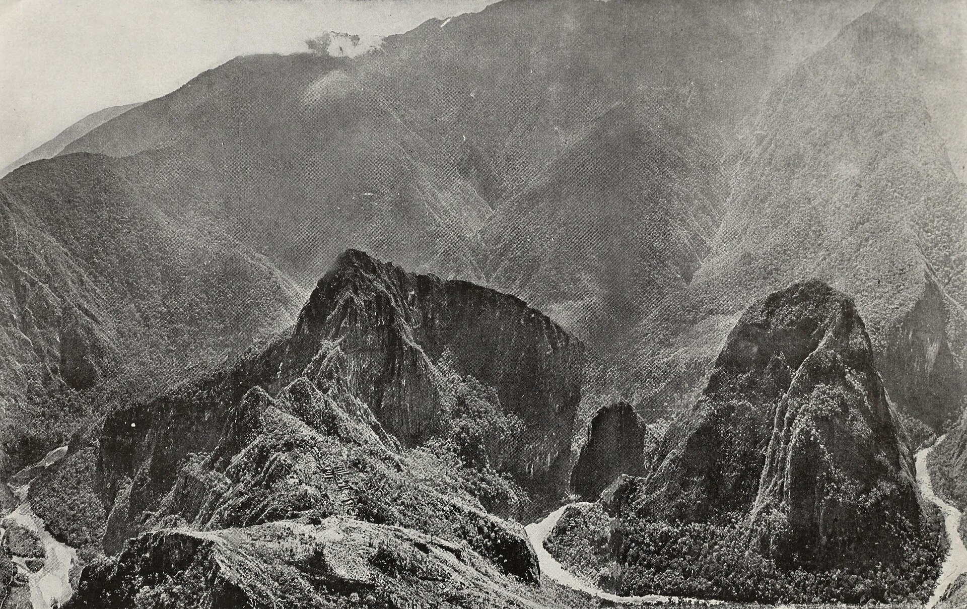 Fotografía antigua en blanco y negro de unas ruinas de piedra sobre una montaña, con un cañón profundo y montañas alrededor