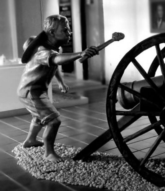 Escultura de Narciso Mendoza: un niño descalzo con sombrero que acerca una mecha encendida a un cañón antiguo