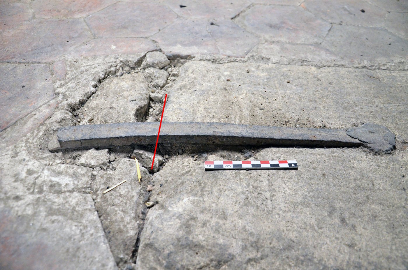 Una barra de hierro oscura y alargada, rota, encajada entre losas de piedra gris, junto a una regla de diez centímetros