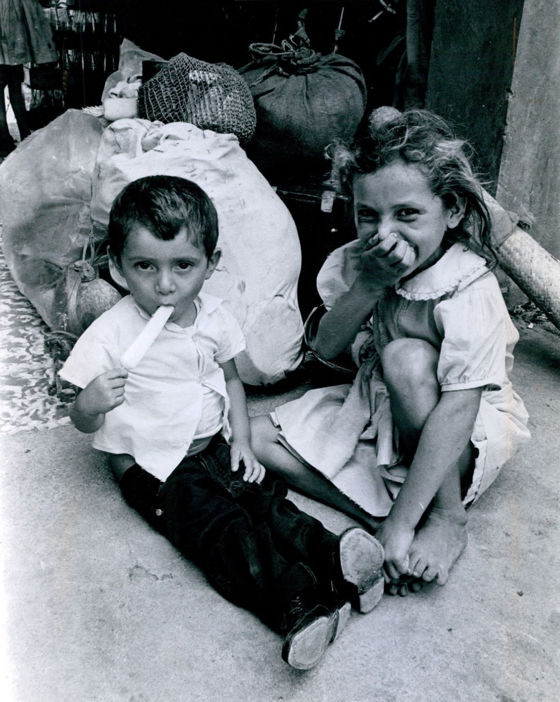Fotografía en blanco y negro de dos niños sentados en el suelo junto a bultos y sacos