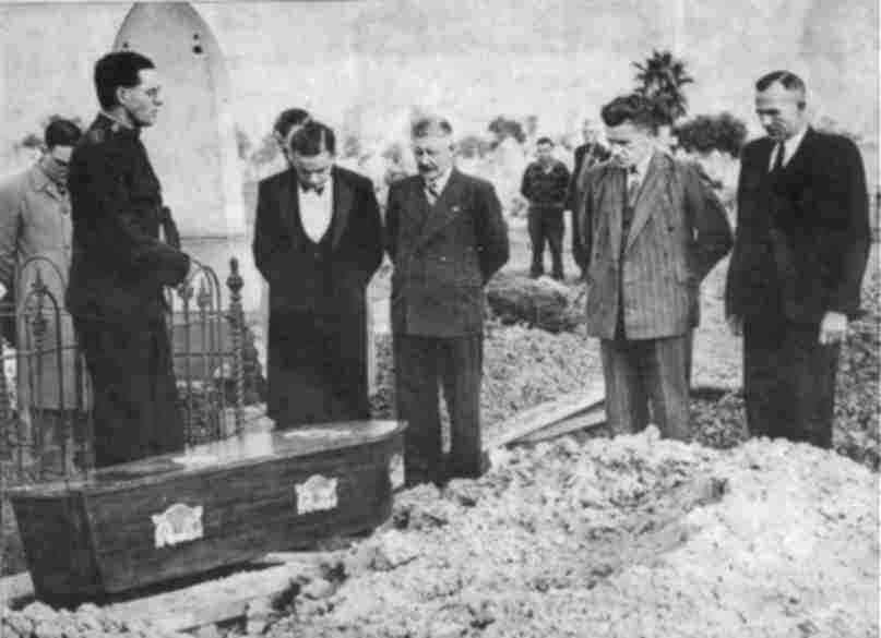 Fotografía en blanco y negro de varios hombres de traje alrededor de un ataúd en un cementerio